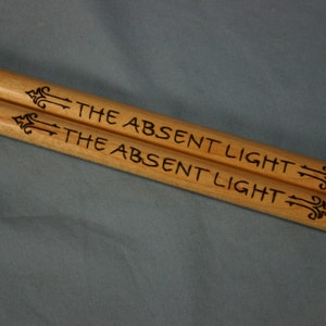 May include: A pair of wooden drumsticks with the words "The Absent Light" engraved on them.