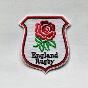 Puede incluir: Parche bordado con el logotipo de la selección inglesa de rugby. El parche tiene un fondo blanco con una rosa roja y hojas verdes en la parte superior. Las palabras "England Rugby" están en texto negro en la parte inferior, y el parche está delineado en rojo.