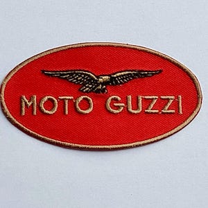 Może przedstawiać: Okrągła czerwona naszywka z napisem "MOTO GUZZI" w złotym kolorze. Szczegółowy złoto-czarny emblemat orła jest umieszczony nad tekstem. Naszywka ma złotą obwódkę.