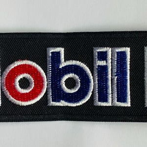 May include: Black rectangular patch with the Mobil 1 logo embroidered in white, red, and blue. The word "Mobil" is in blue with a red "O". The number "1" is in a white square.