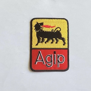 Könnte beinhalten: Gestickter Aufnäher mit dem Agip-Logo. Der obere Teil ist gelb mit einem schwarzen, hundeähnlichen Wesen mit roter Zunge. Der untere Teil ist rot mit dem Wort "Agip" in Weiß.