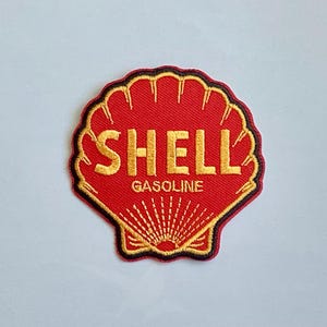 May include: A red, embroidered patch in the shape of a seashell. The patch features the word "SHELL" in gold lettering above "GASOLINE". The shell design is outlined in gold and black.