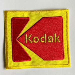 Puede incluir: Parche bordado con el logotipo de Kodak. El parche tiene un borde y un fondo amarillos. Una forma de flecha roja con la palabra "Kodak" en letras doradas está en el centro. El parche es cuadrado y tiene un borde cosido.