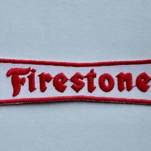 May include: Embroidered patch with the word "Firestone" in red, stylized lettering. The text is on a white background, framed by a red border. The patch has a rectangular shape.