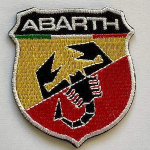 May include: Embroidered patch featuring the word "ABARTH" above a black scorpion emblem. The patch has a shield shape with yellow, red, and green sections, and a black border with white stitching. The patch is likely for automotive enthusiasts.