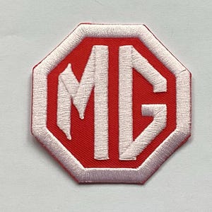 Puede incluir: Parche bordado con el logotipo de MG. El diseño presenta las letras "MG" en blanco sobre un fondo rojo octogonal, con un borde blanco. Un emblema automovilístico clásico.