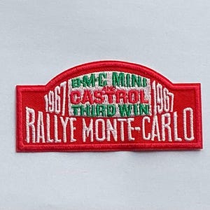 May include: Embroidered red patch with white text, featuring "RALLYE MONTE-CARLO" and "1967". The patch also includes "BMC MINI CASTROL THIRD WIN" in green and white. A vintage-style collectible.