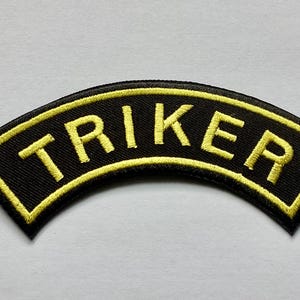 May include: Black, arched patch with the word "TRIKER" embroidered in yellow. The patch has a yellow border and is likely an emblem or insignia.