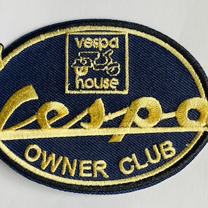 May include: Embroidered patch with a navy blue oval background and gold lettering. The patch features the word "Vespa" in a stylized font, along with the words "Owner Club" and "Vespa House". A gold outline frames the patch.