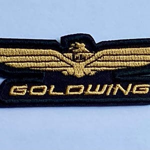 May include: A black and gold embroidered patch featuring a stylized wing design with the word "GOLDWING" in gold lettering. The patch has a detailed, textured appearance, suitable for application on clothing or accessories.