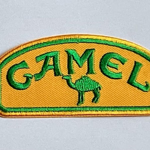 以下が含まれることがあります： 緑色の「CAMEL」の文字と緑色のラクダのシルエットが描かれた刺繍パッチ。黄色い背景に緑色の縁取りと上部の曲線が特徴です。衣類やアクセサリー用のパッチです。