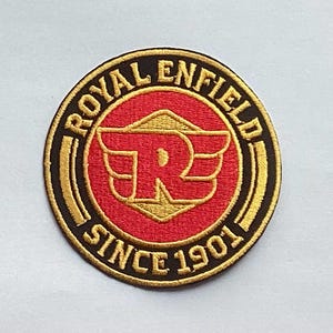 Puede incluir: Parche redondo bordado con las palabras "ROYAL ENFIELD" y "SINCE 1901" en letras doradas. El parche presenta un centro rojo con un emblema dorado de la letra "R" con alas. El borde es negro y dorado.