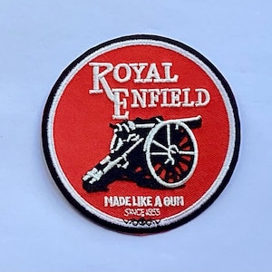 May include: A round patch with a red background and a white border. The text "ROYAL ENFIELD" is arched across the top in white, with a black cannon illustration below. The text "MADE LIKE A GUN SINCE 1955" is at the bottom.