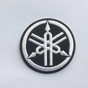 May include: Embroidered patch featuring the Yamaha logo. The patch is circular, with a black background and a white, detailed emblem. The emblem includes a stylized design with arrows and a central vertical element, all outlined in white.