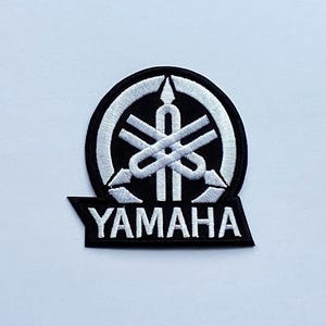 Puede incluir: Parche bordado con el logotipo de Yamaha. El parche tiene un fondo negro con un emblema blanco y la palabra "YAMAHA" en letras blancas. El emblema es un diseño estilizado con formas que se cruzan.