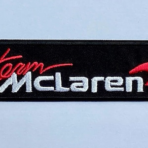 Pode incluir: Patch retangular preto bordado com as palavras "team McLaren" em vermelho e branco. Um design vermelho em forma de swoosh está no lado direito. Ideal para roupas.