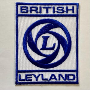 Op de afbeelding: Geborduurde patch met de woorden "BRITISH" en "LEYLAND" in blauw op een witte achtergrond. Het midden heeft een blauw wervelontwerp met de letter "L". De patch is rechthoekig.