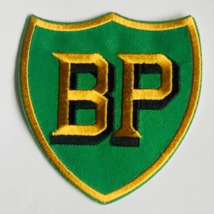 May include: A green shield-shaped patch with gold trim and the letters "BP" in gold with black shadowing. The patch has a textured appearance, suggesting it is embroidered or sewn onto fabric.