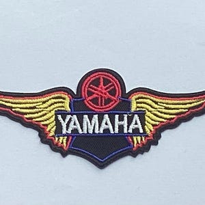 May include: Embroidered Yamaha patch featuring a winged emblem with the brand name. The patch has a black background with red, yellow, and blue accents. The Yamaha logo is in a red circle.