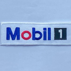 Op de afbeelding: Geborduurde patch met het Mobil 1 logo. Het woord "Mobil" is blauw met een rode "o", en "1" is wit op een zwart vierkant. De patch is rechthoekig met een witte rand, voor kleding of accessoires.