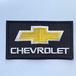 Könnte beinhalten: Schwarzer rechteckiger Aufnäher mit dem Chevrolet-Logo. Das Logo ist eine goldene Fliege mit weißem Rand, über dem weißen Text "CHEVROLET". Dies ist wahrscheinlich ein gesticktes Emblem.