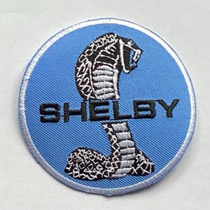 Puede incluir: Un parche redondo bordado con un diseño de cobra en negro, blanco y gris sobre un fondo azul claro. La palabra "SHELBY" está bordada en negro debajo. El parche tiene un borde blanco.