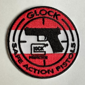 May include: Circular embroidered patch with a red and white striped border. The patch features the word "GLOCK" in black, a black pistol silhouette, and the words "SAFE ACTION PISTOLS". The word "PERFECTION" is also visible.