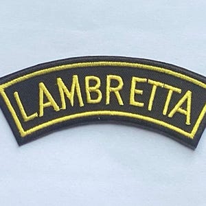 Pode incluir: Um patch preto arqueado com a palavra "LAMBRETTA" bordada em amarelo. O patch tem uma borda amarela, provavelmente um logotipo ou emblema.