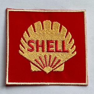 Puede incluir: Parche bordado con el logotipo de Shell. El parche es un cuadrado rojo con un diseño de concha dorada y la palabra "SHELL" en rojo. El parche tiene un borde dorado.