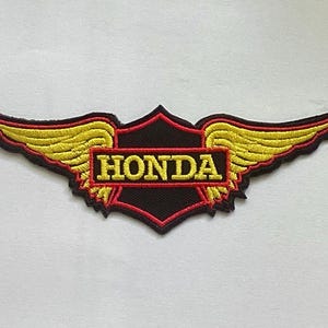 May include: Embroidered Honda patch featuring a black shield with the brand name in yellow. The shield is framed by red and yellow wings, creating a dynamic design. Ideal for jackets, bags, or other apparel.