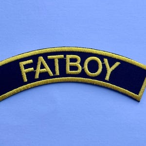 Op de afbeelding: Geborduurde patch met het woord "FATBOY" in gele hoofdletters op een marineblauwe, gebogen rechthoek. De patch heeft een gouden rand en is geplaatst tegen een lichtblauwe achtergrond.