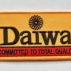Può includere: Toppa ricamata con la parola "Daiwa" in nero, sopra il testo "COMMITTED TO TOTAL QUALITY" in rosso. La toppa ha uno sfondo giallo con un disegno a vortice nero.