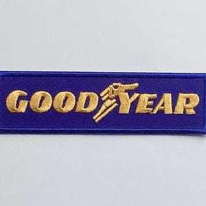 Pode incluir: Um patch retangular com o logotipo Goodyear. O patch tem um fundo azul escuro com as palavras "GOOD YEAR" bordadas em fio dourado. O logotipo Goodyear com o pé alado também é dourado.