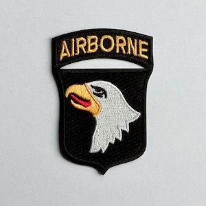 Op de afbeelding: Geborduurde patch met het woord "AIRBORNE" in goud boven een zwart schild. Het schild toont een witte adelaarskop met een gele snavel en rode tong. Geschikt voor militaire of luchtvaartliefhebbers, een symbool van luchtlandingstroepen.