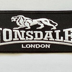 May include: Black rectangular patch featuring white embroidered text and a lion emblem. The text reads "LONSDALE LONDON". This patch is likely designed for clothing or accessories.