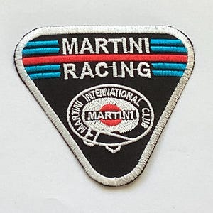 Puede incluir: Parche triangular negro con las palabras blancas "MARTINI RACING". El parche presenta rayas azules y rojas, y un emblema circular con "MARTINI INTERNATIONAL CLUB" y el logotipo de Martini.