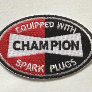 May include: Embroidered Champion Spark Plugs patch. The oval patch features a red and black split design with the words "Equipped With Champion Spark Plugs" and "Champion" in white lettering. The patch has a black border.