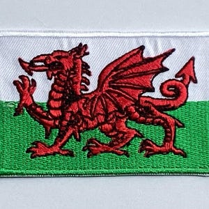 May include: Embroidered patch of the Welsh flag. It features a red dragon on a white and green background. The dragon is depicted with wings and a curled tail. The patch is rectangular with a stitched border.