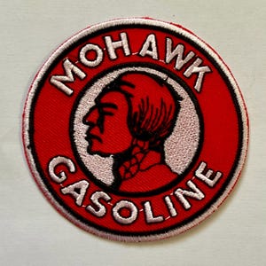 Op de afbeelding: Een ronde, rode patch met de woorden "MOHAWK GASOLINE" in witte letters. De patch toont een profiel van een Indiaanse man in een witte cirkel. Het profiel van de man is rood, met een zwarte vlecht. De patch heeft een zwarte rand.