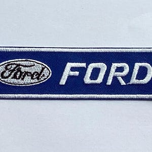 Puede incluir: Un parche rectangular azul bordado con un borde blanco. El parche presenta el logotipo de Ford en un óvalo blanco y la palabra "FORD" en letras mayúsculas blancas.