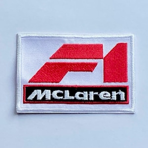 May include: Embroidered patch with the McLaren logo. The patch has a white background with a red and black logo. The McLaren logo is a stylized red "F1" design above the word "McLaren" in white.