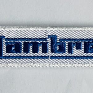 May include: Embroidered patch featuring the word "Lambretta" in blue, with a blue, white, and red bullseye design on the left side. The patch is rectangular with a white background and a stitched border.