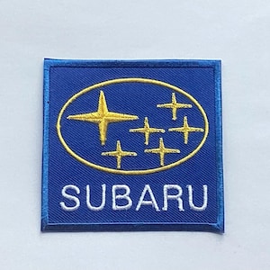 Puede incluir: Parche bordado azul cuadrado con el logotipo de Subaru. El logotipo incluye un óvalo dorado con una estrella dorada y cuatro estrellas doradas más pequeñas. La palabra "SUBARU" está bordada en blanco debajo.