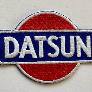 May include: Embroidered patch featuring the word "DATSUN" in white, stitched onto a blue rectangular background. The rectangle is set against a red, semi-circular backdrop, all outlined with white stitching. A vintage-style emblem.