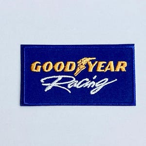 以下が含まれることがあります： 青い長方形の背景に、金と白で「GOOD YEAR Racing」と刺繍されたパッチ。グッドイヤーのロゴである翼の生えた足も金色です。このパッチはおそらく衣類や収集品用です。
