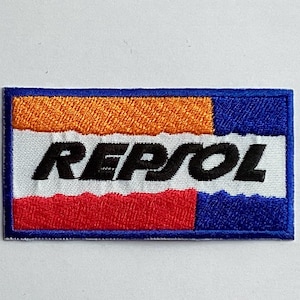 May include: Embroidered patch featuring the word "REPSOL" in black, set against a white background. The patch has a rectangular shape with blue borders and sections of orange and red. The design is a logo.