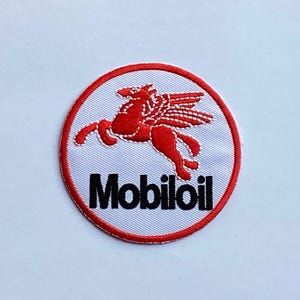 Pode incluir: Patch bordado circular com o logotipo da Mobil Oil. O patch tem uma borda vermelha e um fundo branco. Um Pégaso vermelho com asas está no centro, acima do texto preto "Mobiloil".