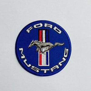 Könnte beinhalten: Ein runder, blauer, gestickter Aufnäher mit dem Ford Mustang-Logo. Das Logo zeigt ein silbernes, galoppierendes Pferd über einem vertikalen Streifen in Rot, Weiß und Blau. Die Wörter "FORD" und "MUSTANG" sind weiß gestickt.