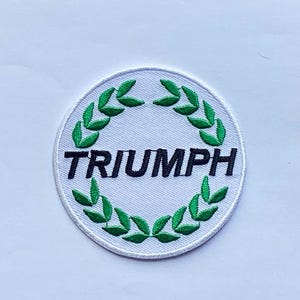 May include: Embroidered patch with the word "TRIUMPH" in black, surrounded by a green laurel wreath on a white background. The patch has a white border and is circular in shape.