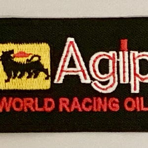 May include: Black rectangular patch with embroidered text and graphics. Features the word "Agip" in white with red accents, and "WORLD RACING OILS" in red. Includes a yellow square with a black silhouette of a dog-like creature.